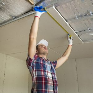 Young handsome man in casual clothing takes measurement of drywall suspended ceiling connected to metal frame on ceiling insulated with aluminum foil. Construction, renovation, DIT concept.